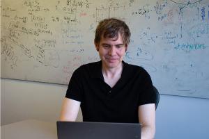 A man sits at a laptop, in front of a dry erase board covered in complicated equations.
